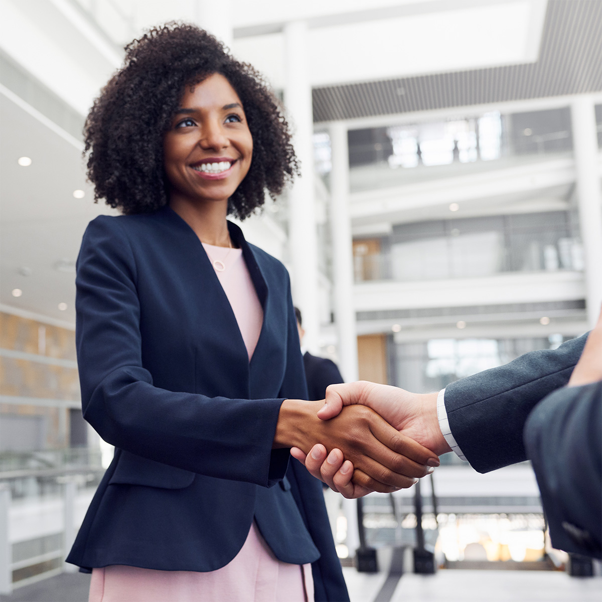 A women in a suit jacket shaking hands with someone in a brightly lit office building.